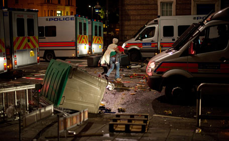 A woman walks through the debris with two children as riot police try to contain a large group of people on a main road in Tottenham, north London on August 6, 2011. Two police cars were on Saturday set ablaze in north London following a protest over the fatal shooting of a 29-year-old man in an armed stand-off with officers. The patrol cars were torched as dozens gathered outside the police station on the High Road in Tottenham. (AFP)
