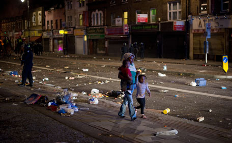 A woman walks through the debris with two children as riot police try to contain a large group of people on a main road in Tottenham, north London on August 6 2011.  Two police cars were on Saturday set ablaze in north London following a protest over the fatal shooting of a 29-year-old man in an armed stand-off with officers.  The patrol cars were torched as dozens gathered outside the police station on the High Road in Tottenham. (AFP)
