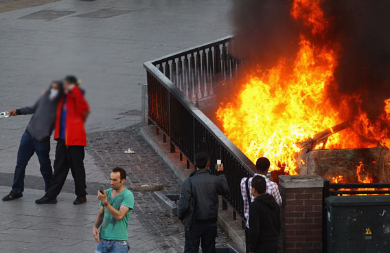 People take photographs of a burning car during riots in Birmingham City Centre in Birmingham, England. After three nights of rioting and looting in and around London, the chaos is starting to spread to other cities around Britain. (GETTY/GALLO)